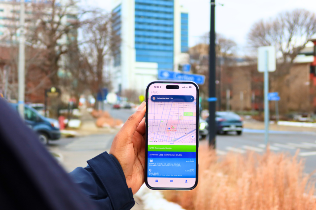 The image shows a person holding a cellphone with the All Access App open. In the background is Buffalo General Hospital.