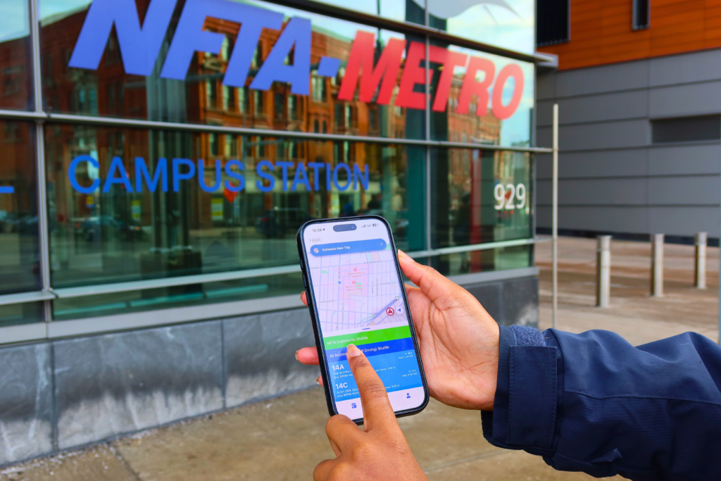 The image shows the hand of a person with the All Access app open on a cellphone. In the background is the NFTA Metro Station at Allen Street in Buffalo.